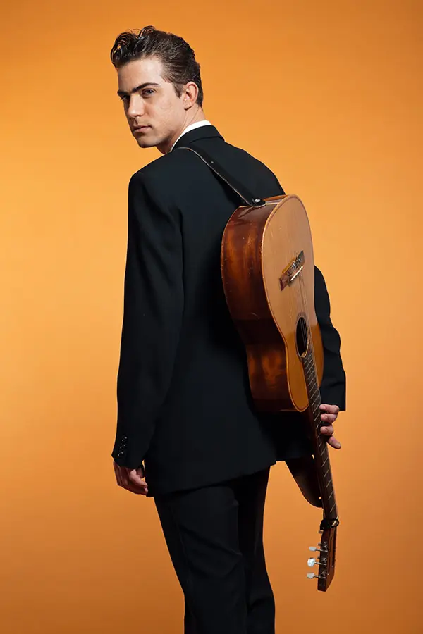 Man in a suit carrying an acoustic guitar on his back