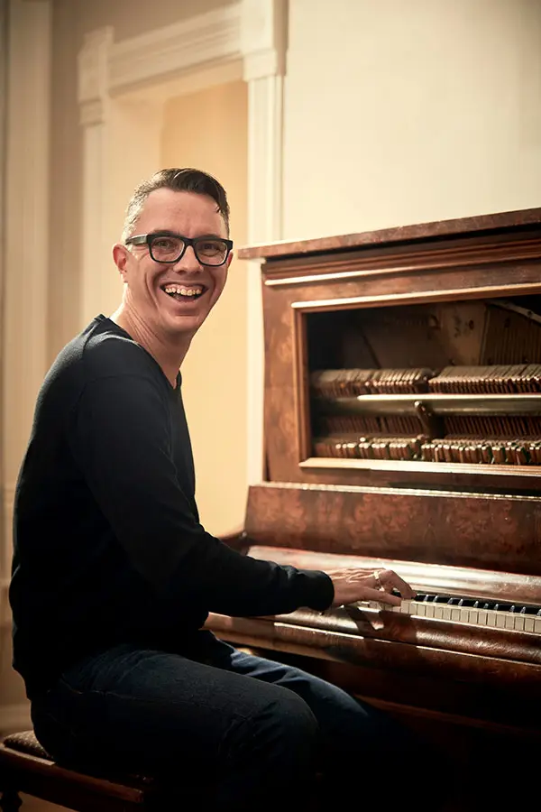 Smiling man with glasses playing an open upright piano