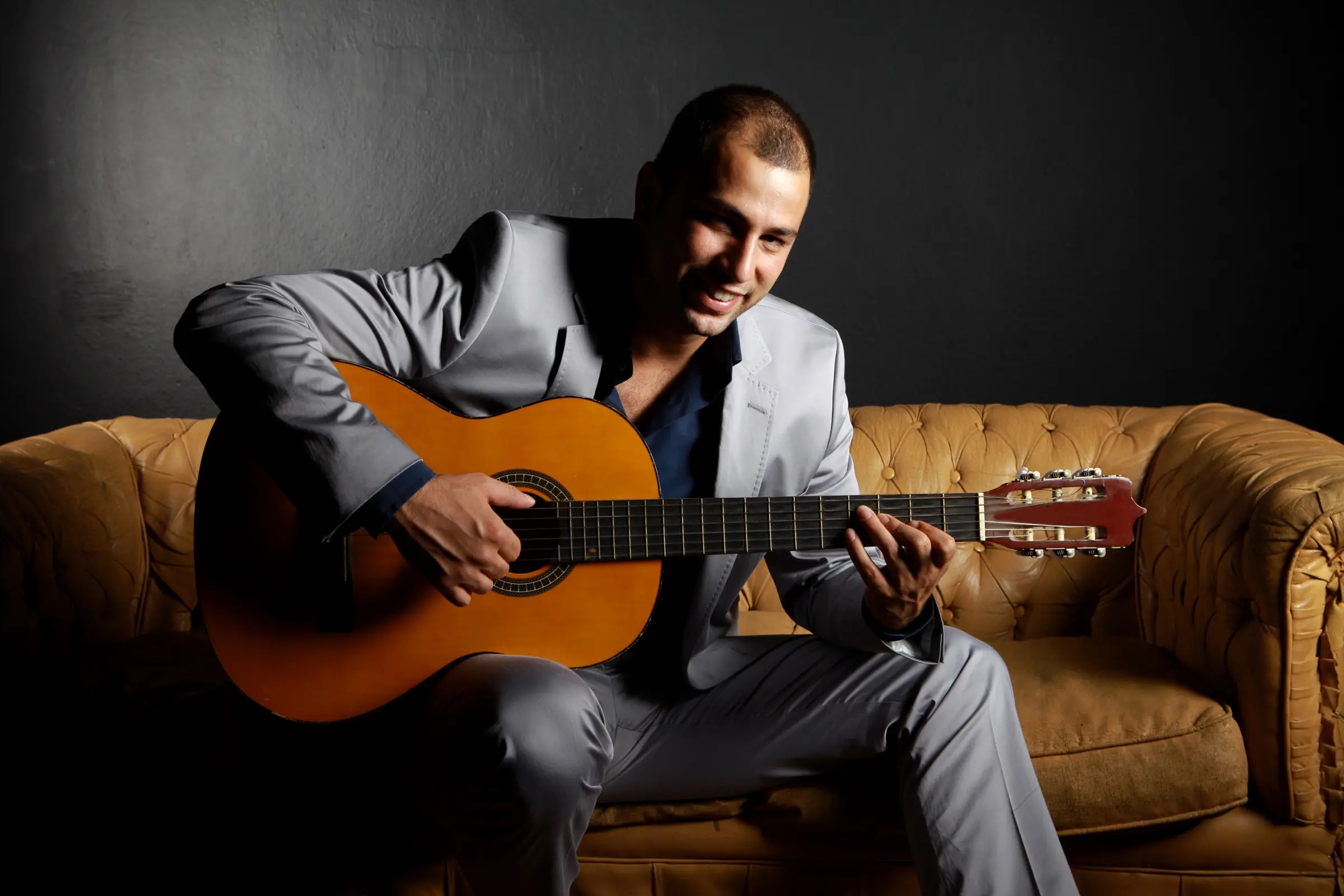 Smiling man in a suit playing a classical guitar on a leather sofa.