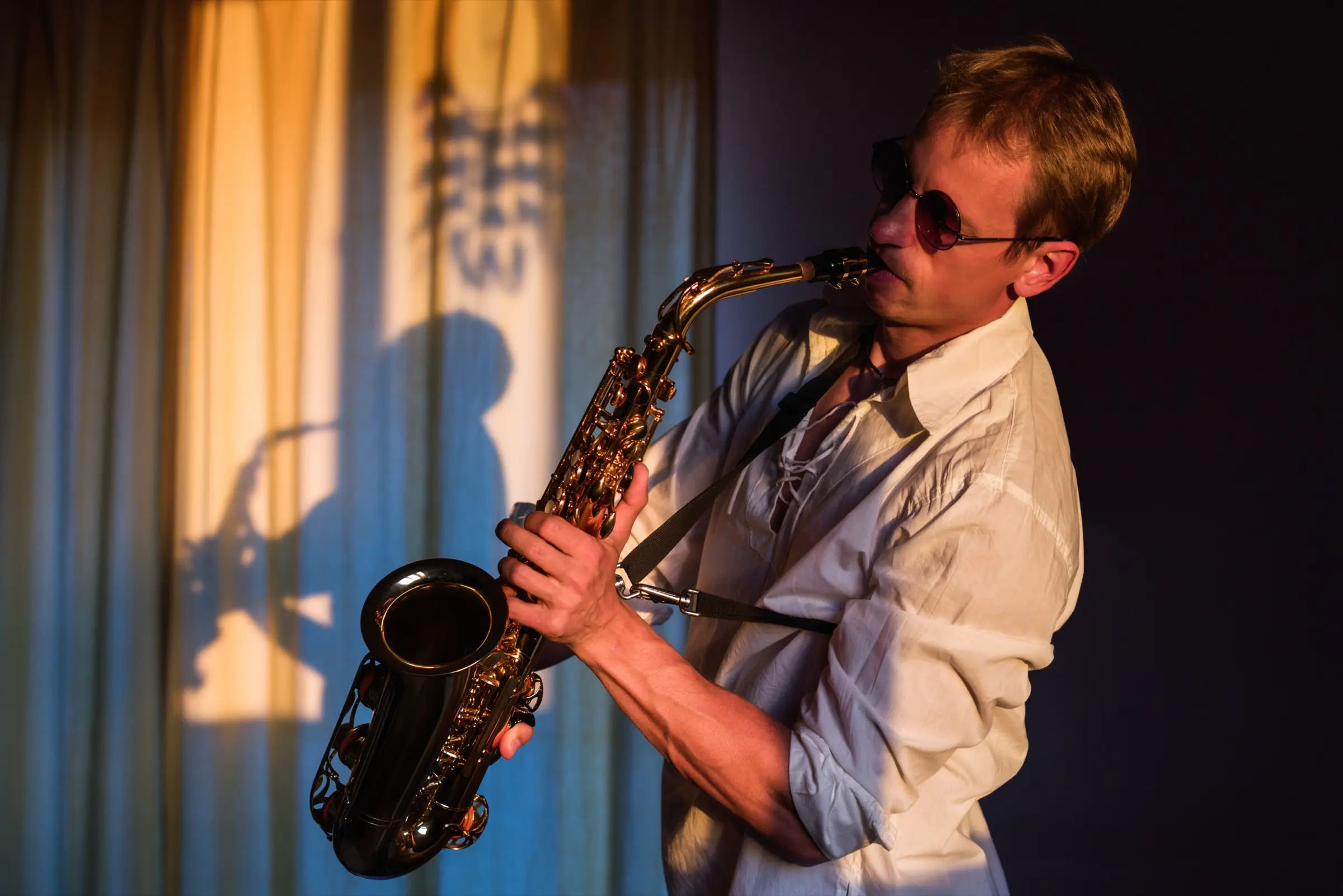 Man in sunglasses playing the saxophone by a curtained window.
