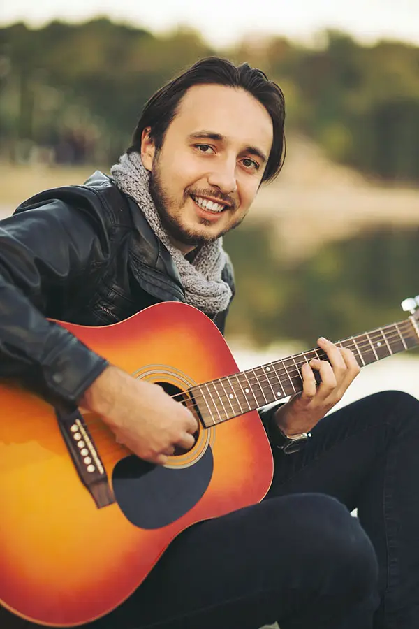 Man sitting outdoors by water playing an acoustic guitar