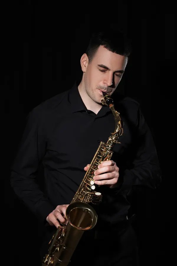 Man in a black shirt playing alto saxophone before a dark curtain