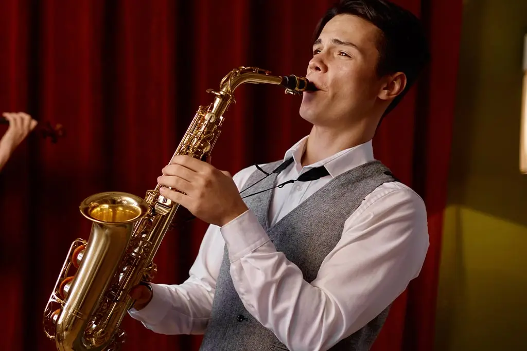 Man in a grey waistcoat playing alto saxophone before red curtains