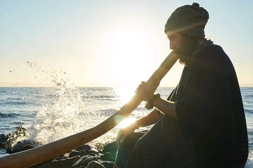 Man playing a didgeridoo on rocks beside the sea