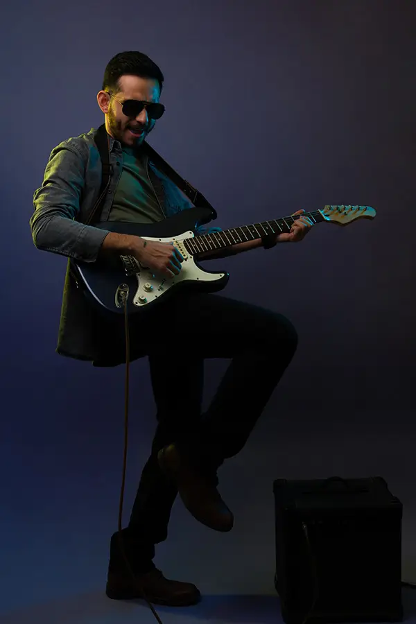 Man wearing sunglasses playing an electric guitar beside an amplifier