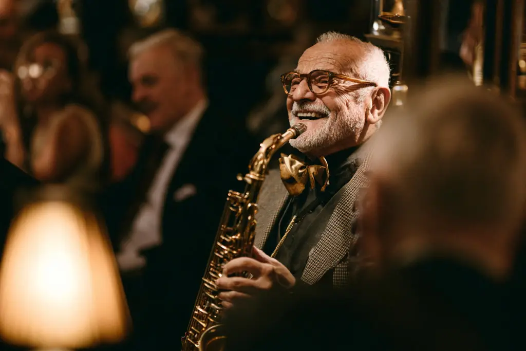 Smiling older man in a bow tie holding a saxophone in a warmly lit bar.