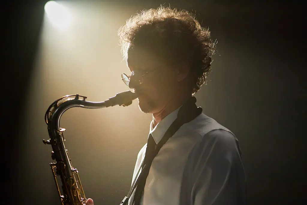 Older man with glasses playing saxophone under stage lighting