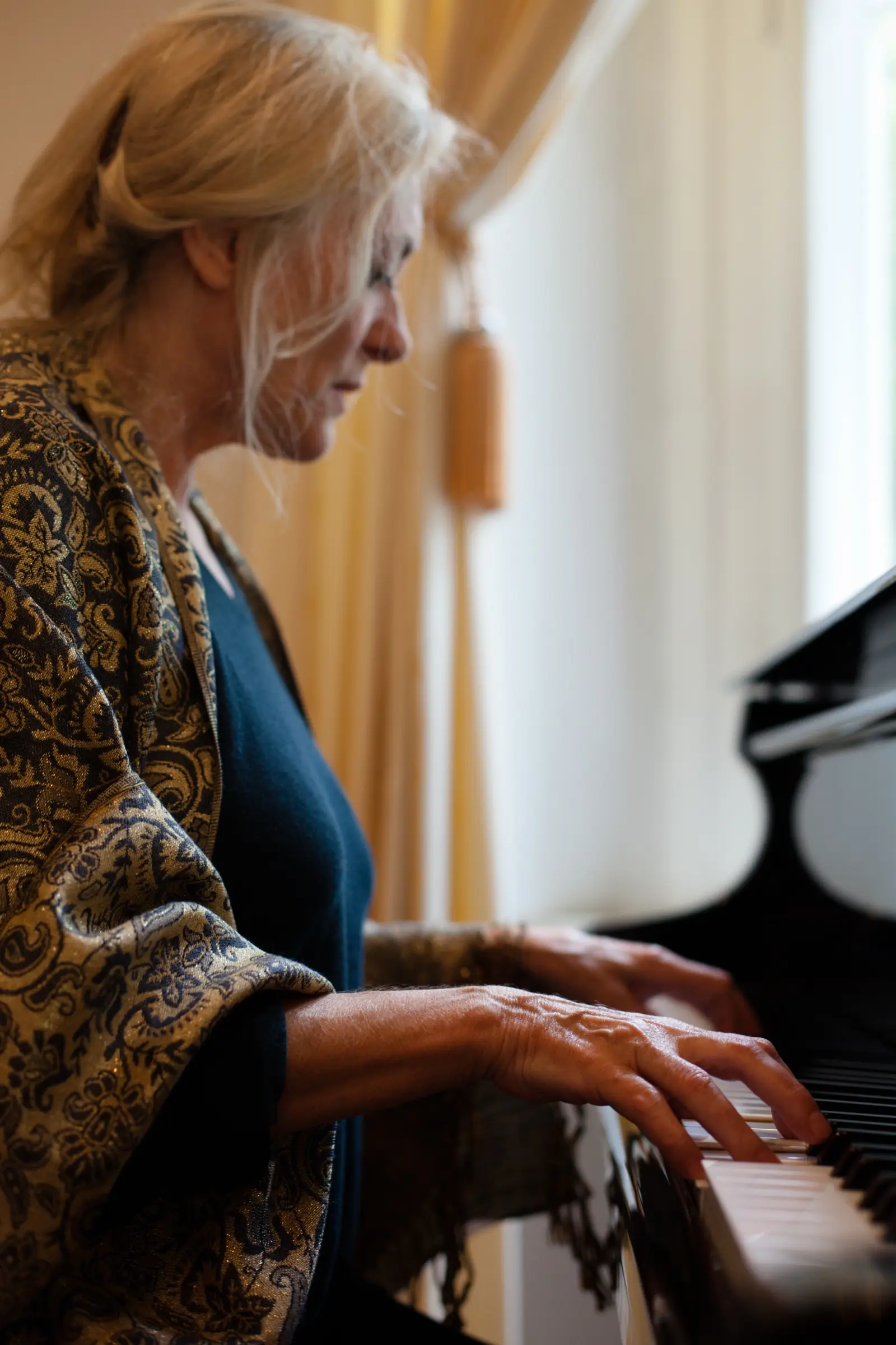 Older woman playing the piano by a curtained window.