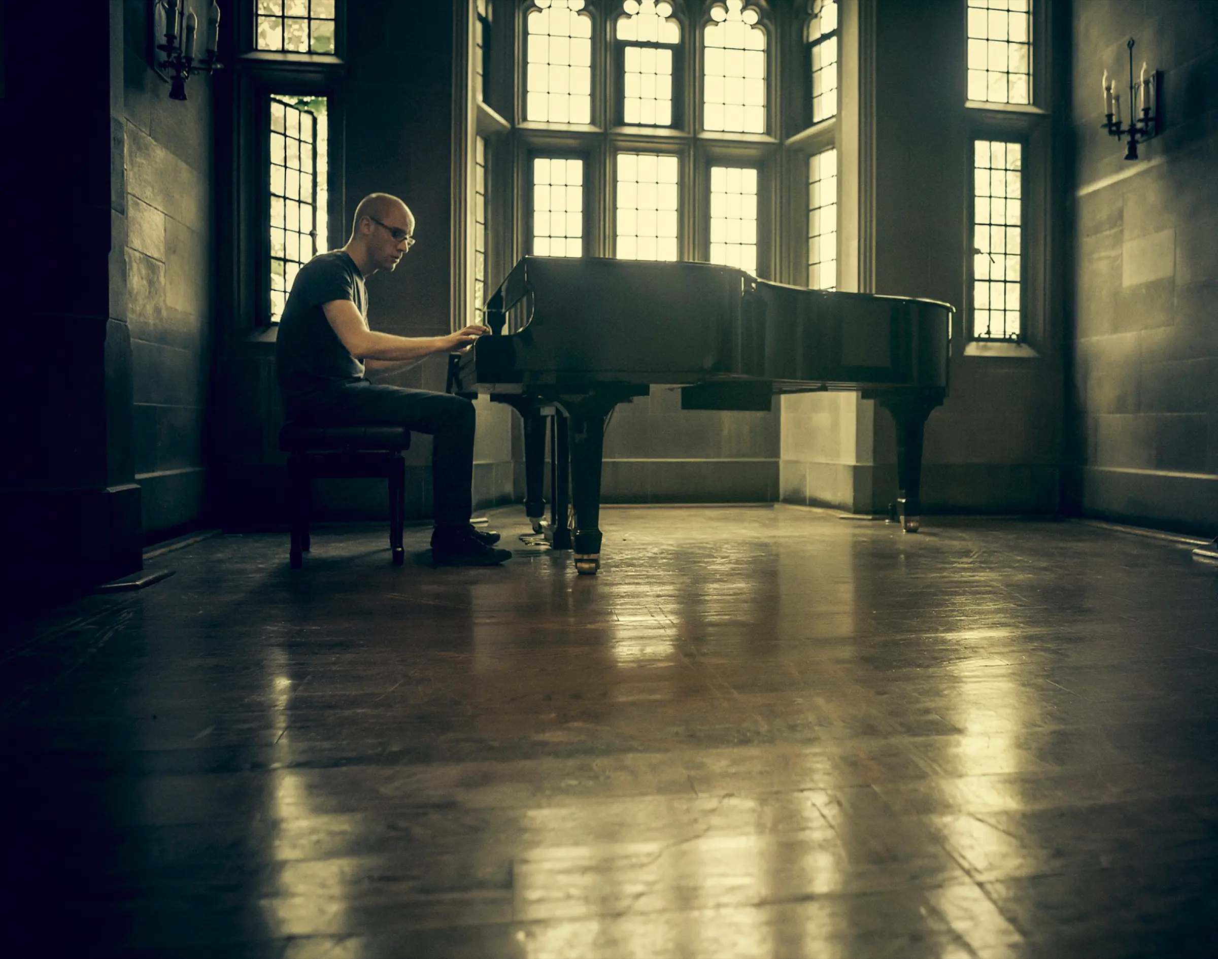 Pianist playing a grand piano alone in a large wood-floored hall.