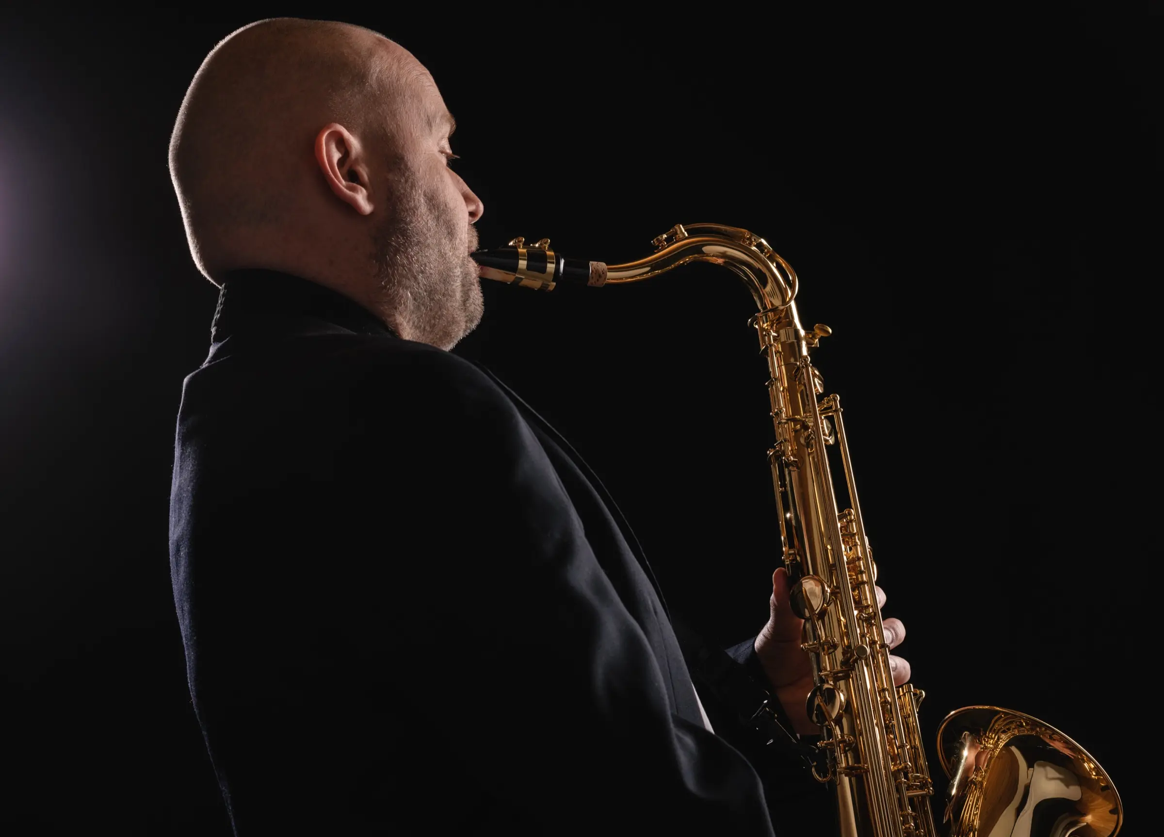 Bald saxophonist playing in profile against a dark background.