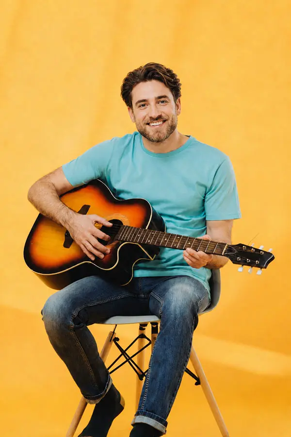 Smiling man in a turquoise shirt with an acoustic guitar on a yellow background