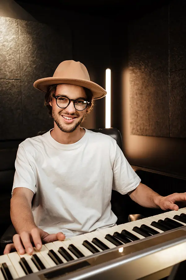 Smiling man in a beige hat playing keyboard in a studio