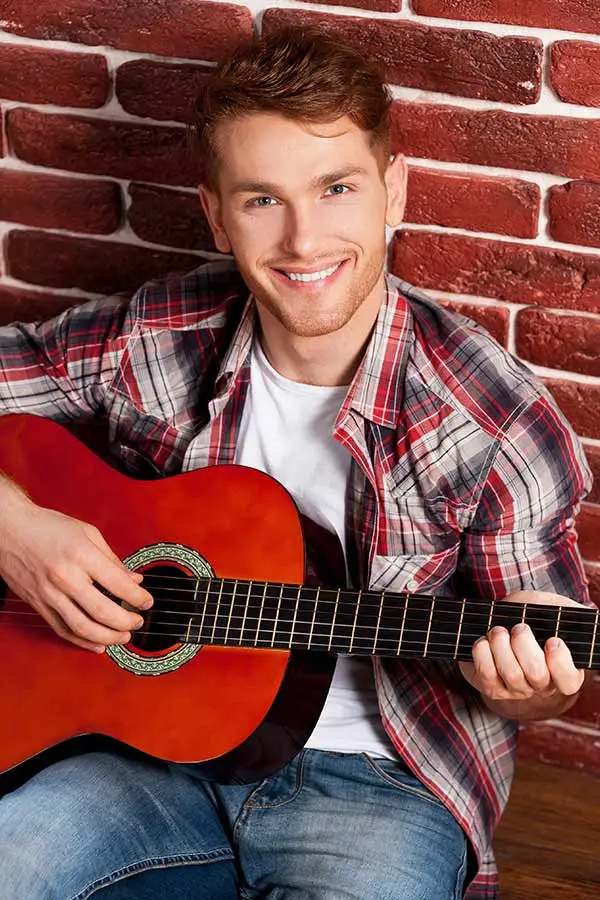 Smiling man in a plaid shirt playing classical guitar by a brick wall