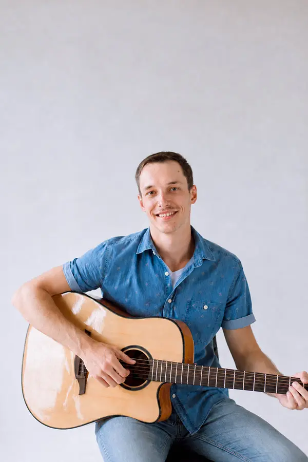 Smiling man in a denim shirt seated with an acoustic guitar