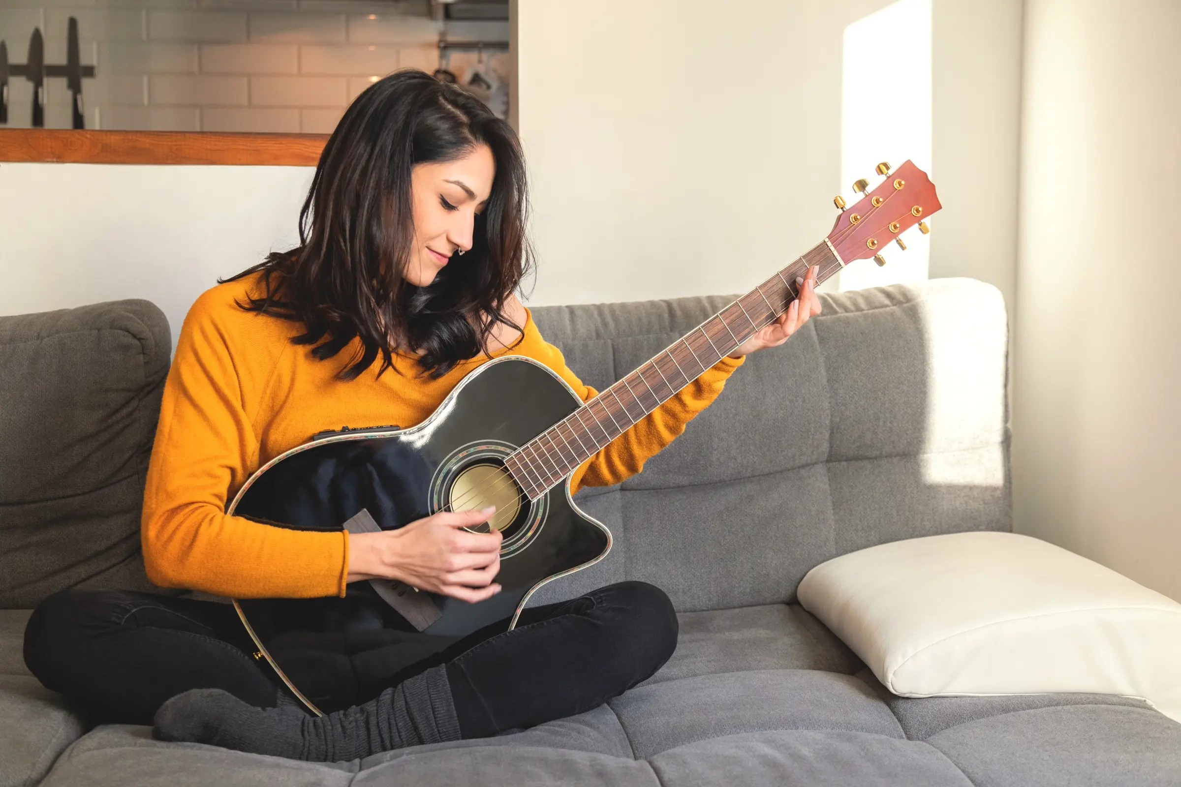 Woman playing an acoustic guitar on a grey sofa.