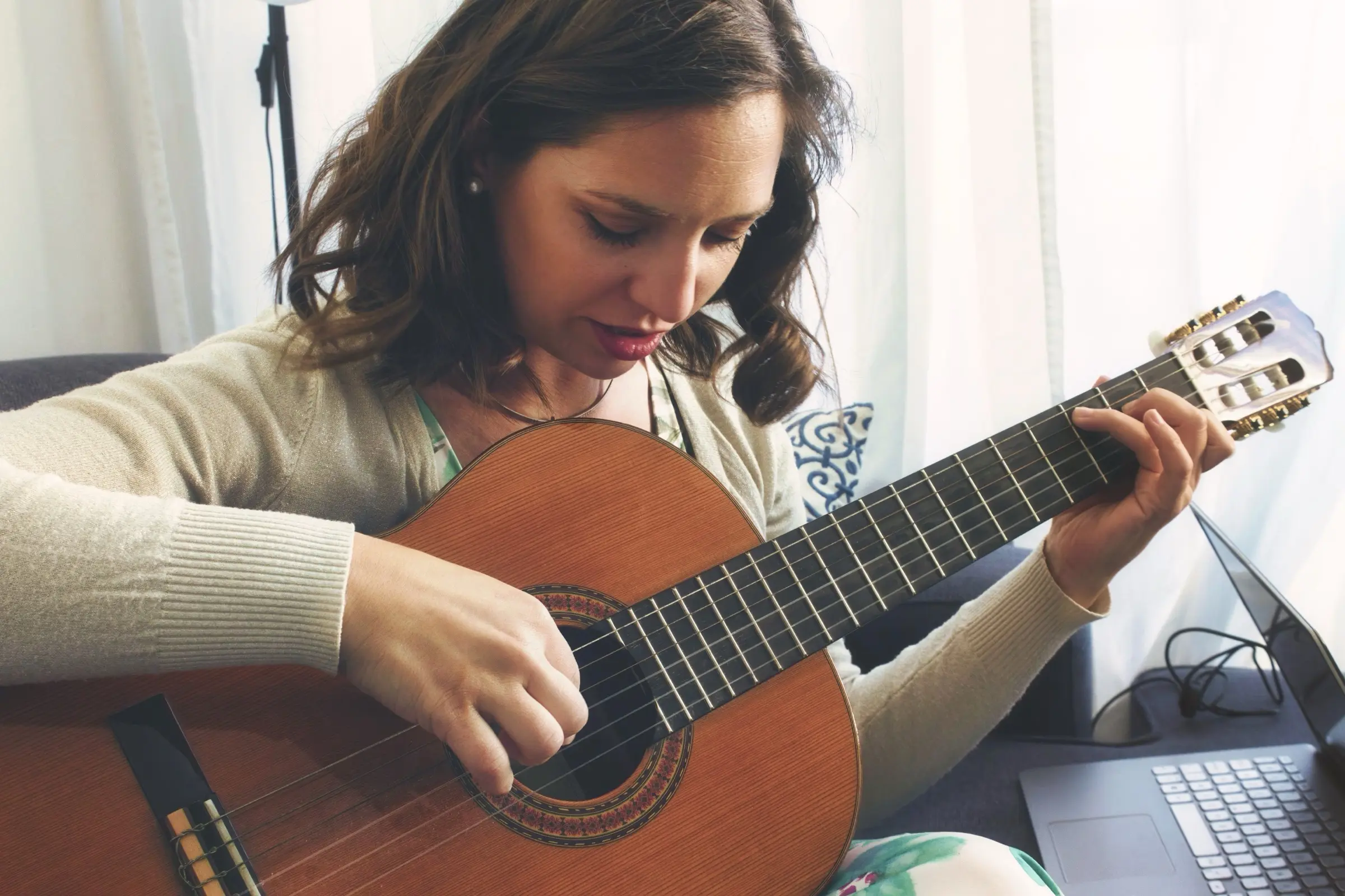 Woman playing a classical guitar beside a laptop.