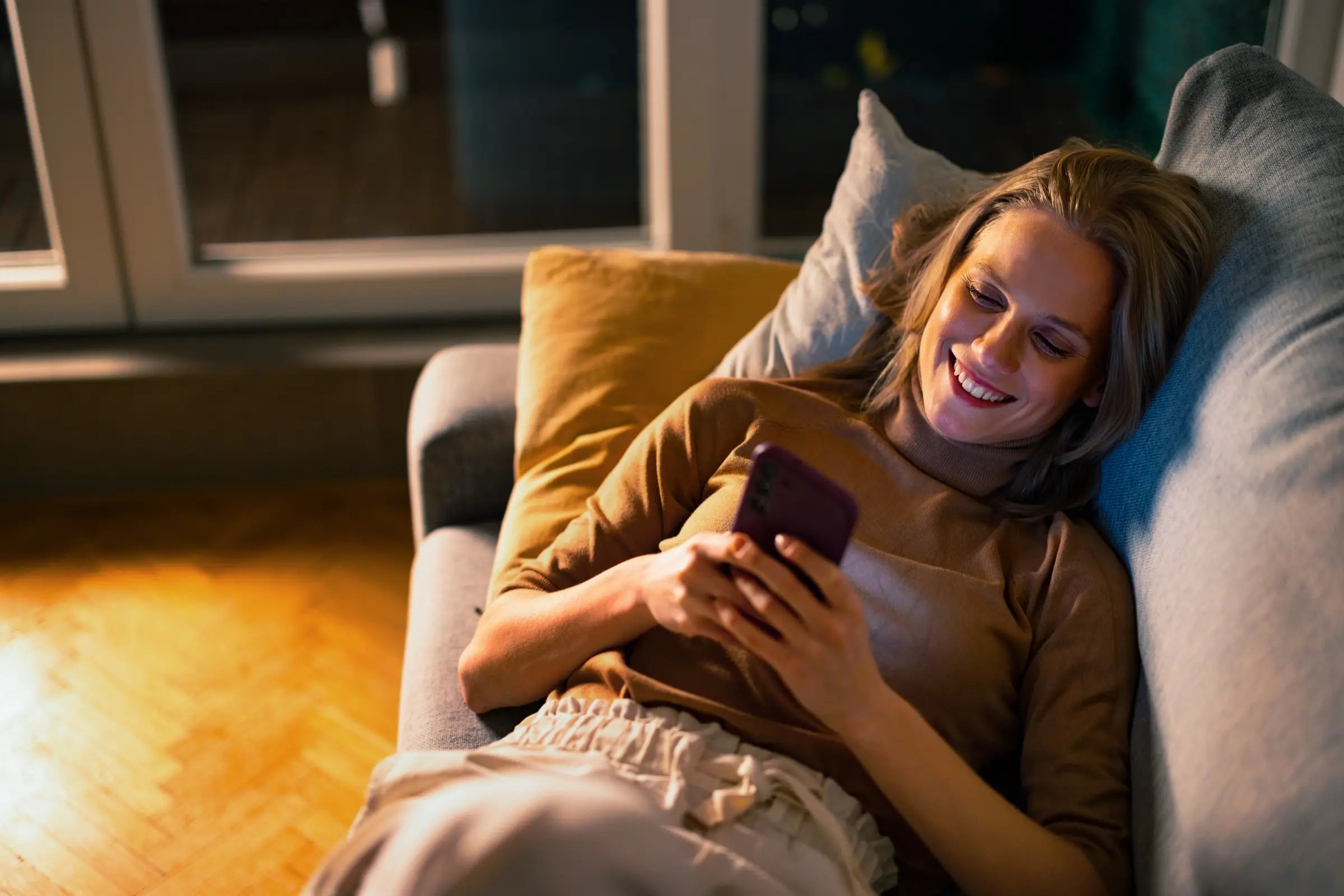 Smiling woman reading a message on her phone on a sofa in the evening