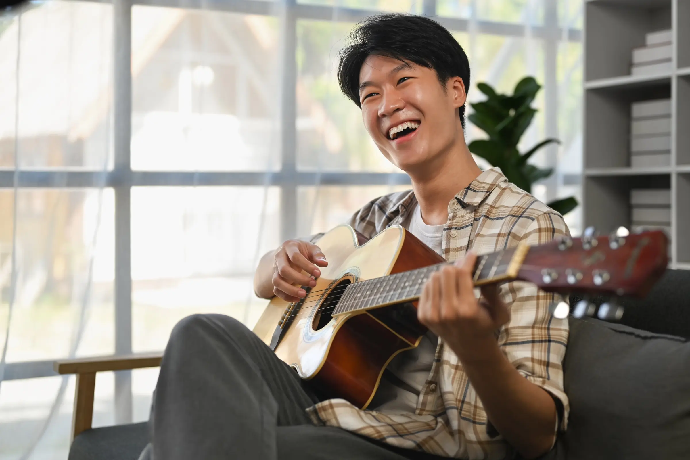 Young man laughing as he plays an acoustic guitar on a sofa.