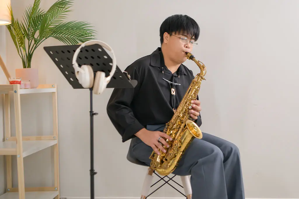 Young man seated beside a music stand playing an alto saxophone